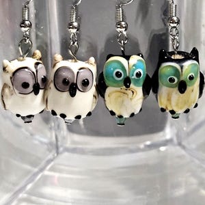 May include: Four pairs of owl-shaped earrings with silver-colored hooks. The owls are white with black and gray or blue and green accents. The earrings are hanging in front of a clear, textured background.