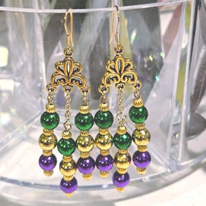 May include: Gold-tone chandelier earrings with green, gold, and purple beads. The earrings feature an ornate filigree design at the top and are suspended from gold-colored ear wires. The beads are arranged in a cascading pattern.
