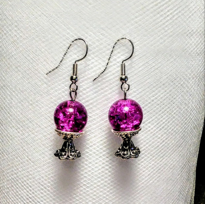 May include: A pair of dangle earrings with a silver base and a round, purple glass bead. The beads have a sparkly, iridescent finish.