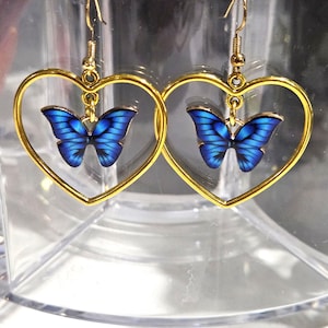 May include: A pair of gold-colored heart-shaped earrings. Each earring features a blue butterfly pendant suspended within the heart frame. The butterfly has detailed wing patterns. The earrings are designed with a hook closure.