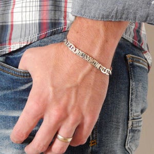 Op de afbeelding: Een zilveren armband met de naam "BENJAMIN" gegraveerd. De armband wordt om een pols gedragen, met een gouden ring om de vinger. De persoon draagt een geruit overhemd en een blauwe spijkerbroek.