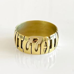 Puede incluir: Un anillo de tono dorado pulido que presenta un diseño de banda ancha con letras recortadas que forman la palabra "IGGAT". El anillo tiene una superficie metálica lisa y reflectante y una forma circular continua de altura uniforme.