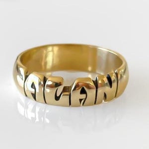 Puede incluir: Un anillo de tono dorado pulido que presenta el nombre ALANI formado por la propia banda de metal. Las letras están integradas en la estructura circular del anillo, creando un diseño metálico sólido y continuo con un acabado de superficie liso y brillante.