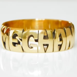 May include: A polished gold-tone ring featuring the name MEGHAN formed by cut-out capital letters that wrap around the entire band. The metal surface is smooth and reflective, creating a continuous design where each letter connects to the next.