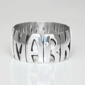 May include: A silver ring with the name "MARK" spelled out in bold, raised letters.
