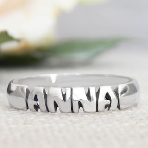 May include: A polished silver-toned metal ring featuring the name ANNA cut out in capital block letters. The band is smooth and rounded, with the letters forming the central design element of the ring, creating a continuous metallic surface.