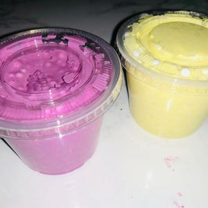 May include: Two clear plastic containers filled with slime. One container is filled with purple slime and the other is filled with yellow slime.