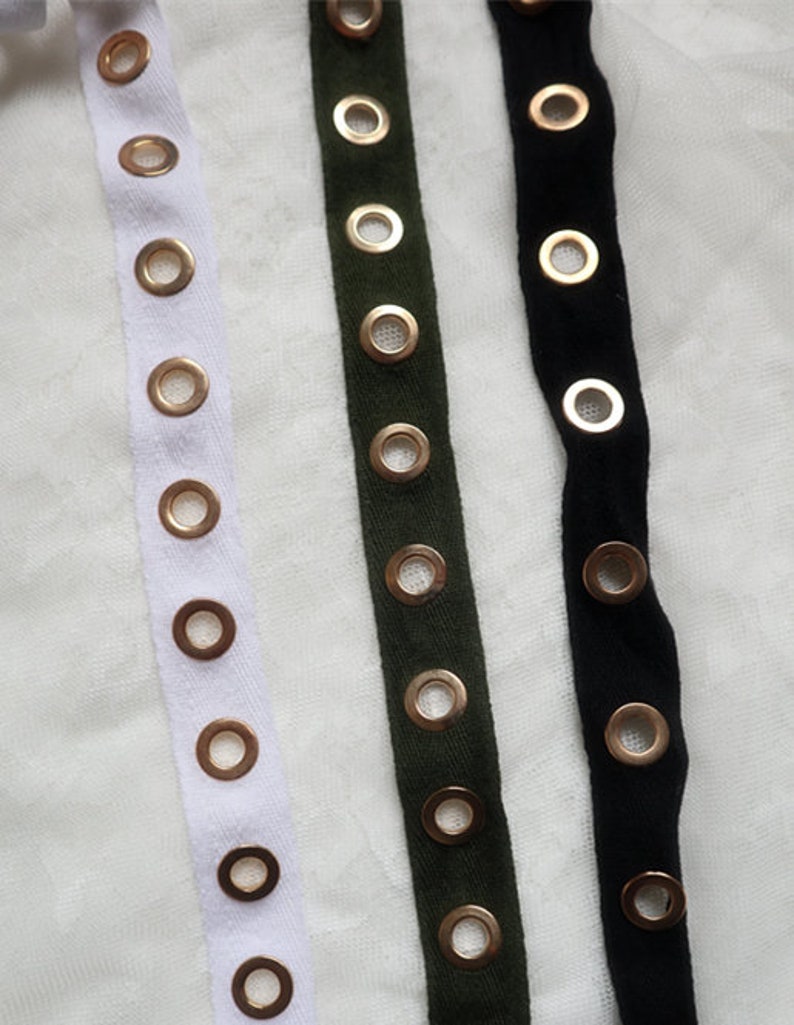 May include: Close-up of three fabric strips: white, olive green, and black. Each strip is adorned with evenly spaced, gold-colored eyelets. The eyelets provide a decorative and functional element, suitable for various crafting and design projects.