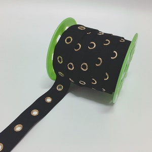 May include: Black ribbon with gold eyelets, rolled on a green spool. The ribbon is made of a soft material and is suitable for various craft projects. The eyelets are evenly spaced along the length of the ribbon.