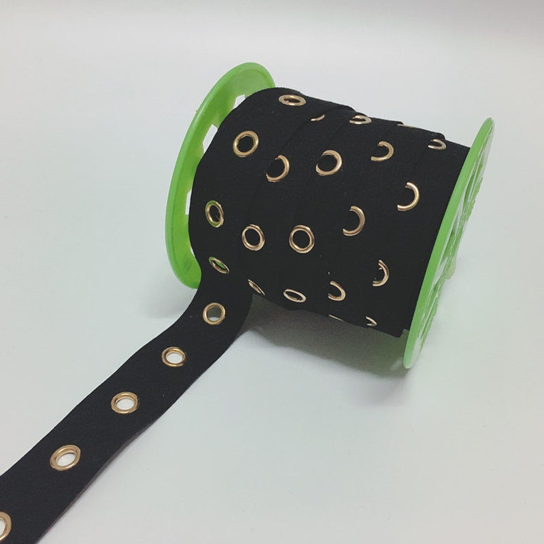 May include: Black ribbon with gold eyelets, rolled on a green spool. The ribbon is made of a soft material and is suitable for various craft projects. The eyelets are evenly spaced along the length of the ribbon.