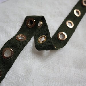 May include: Close-up of a dark green fabric strip with evenly spaced gold eyelets. The fabric is folded and lies on a white, textured surface. Another white strip with gold eyelets is visible in the background.