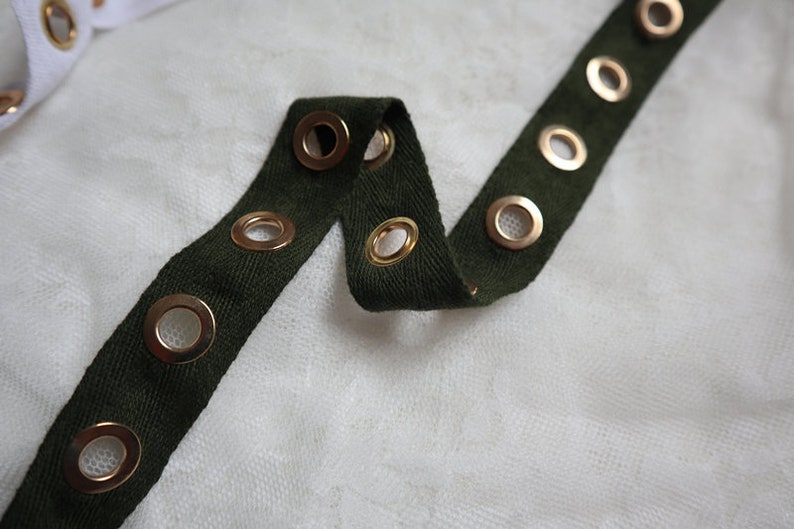 May include: Close-up of a dark green fabric strip with evenly spaced gold eyelets. The fabric is folded and lies on a white, textured surface. Another white strip with gold eyelets is visible in the background.