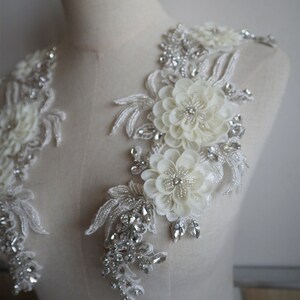 May include: Two white floral appliques with pearl and rhinestone embellishments. The appliques are perfect for adding a touch of elegance to wedding dresses, bridal accessories, or other special occasion garments.