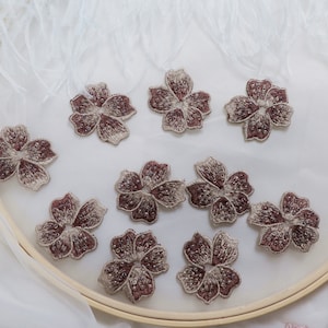 Puede incluir: Primer plano de varios apliques de flores bordadas. Las flores tienen un centro burdeos y pétalos con un contorno plateado-dorado. Los pétalos están detallados con pequeñas cuentas brillantes. Los apliques están dispuestos sobre una superficie blanca.