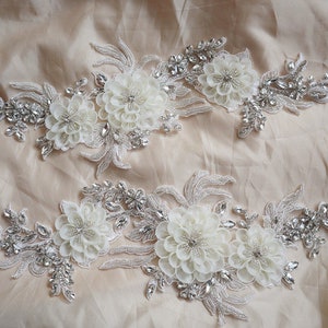 May include: Two white floral appliques with silver beading and rhinestones. The appliques are perfect for adding a touch of elegance to any garment.