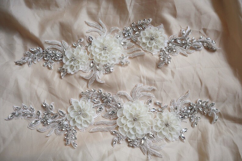 May include: Two white floral appliques with silver beading and rhinestones. The appliques are perfect for adding a touch of elegance to any garment.
