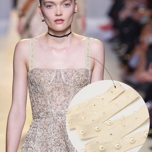 May include: Close-up of a beige fabric strap with white pearl embellishments. The strap is used for bra straps and can be adjusted to fit different sizes. The text "使用方法は: 可以做肩带" is visible in the image.