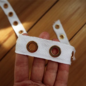 May include: Close-up of white fabric tape with evenly spaced gold eyelets. The tape is held in a hand, with additional sections of the tape visible in the background. The eyelets are circular and metallic.