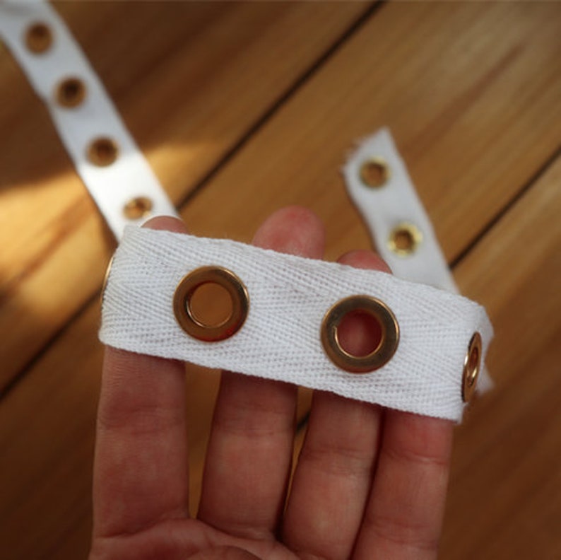 May include: Close-up of white fabric tape with evenly spaced gold eyelets. The tape is held in a hand, with additional sections of the tape visible in the background. The eyelets are circular and metallic.
