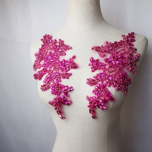 May include: Two fuchsia-colored floral appliques with intricate bead and crystal embellishments. The appliques are arranged symmetrically, featuring a delicate, flowing design. The embellishments are a mix of shapes and sizes, creating a sparkling effect.