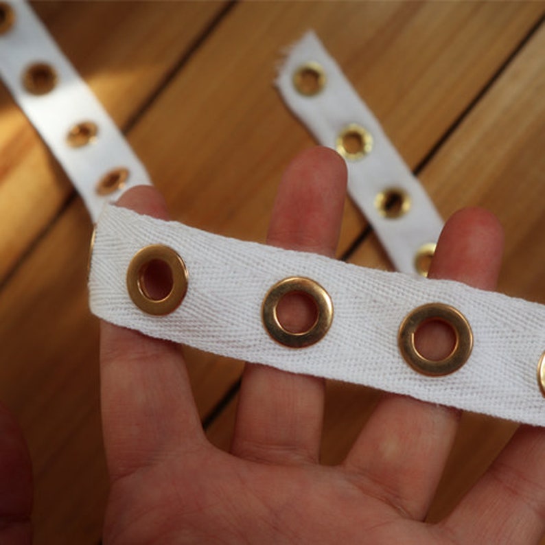 May include: Close-up of white fabric strips with evenly spaced gold eyelets. The strips are held in a hand, with a wooden surface in the background. The eyelets are circular and metallic, creating a decorative element.