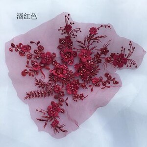 以下が含まれることがあります： ビーズのディテールが付いた、ワインレッドのフラワーモチーフのアップリケ。アップリケは薄いピンクの生地に施されています。