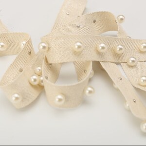May include: Beige fabric ribbon with a herringbone pattern and white pearl beads sewn along the edges.