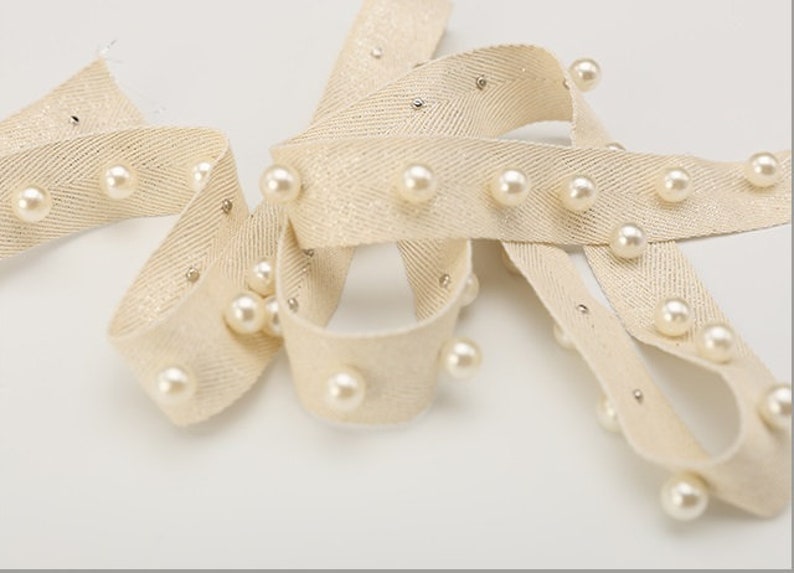 May include: Beige fabric ribbon with a herringbone pattern and white pearl beads sewn along the edges.
