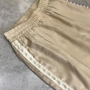May include: A pair of beige pants with a white beaded trim along the side seams. The pants have an elastic waistband.