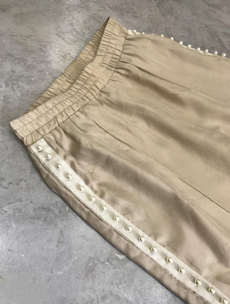 May include: A pair of beige pants with a white beaded trim along the side seams. The pants have an elastic waistband.