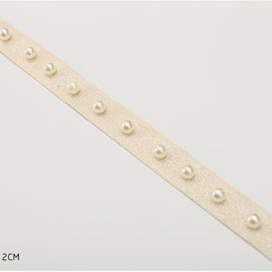 May include: A close-up of a beige ribbon with white pearl embellishments. The ribbon is 2 cm wide.