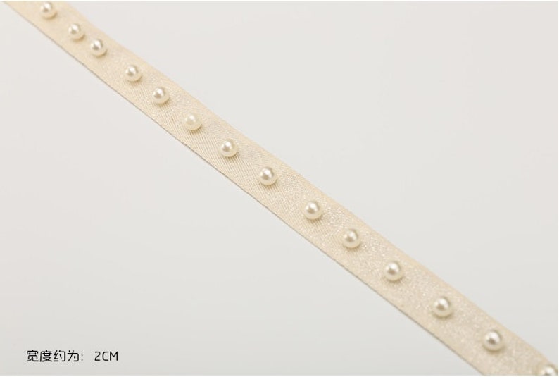May include: A close-up of a beige ribbon with white pearl embellishments. The ribbon is 2 cm wide.
