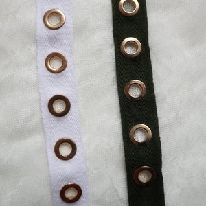 May include: Close-up of two strips of fabric with metal eyelets. One strip is white, the other is dark green. The eyelets are gold-colored and evenly spaced along the fabric strips. The background is a textured white surface.