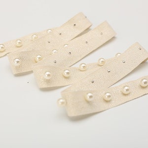 May include: Beige ribbon trim with white pearl embellishments. The ribbon has a herringbone weave and is decorated with evenly spaced pearls.