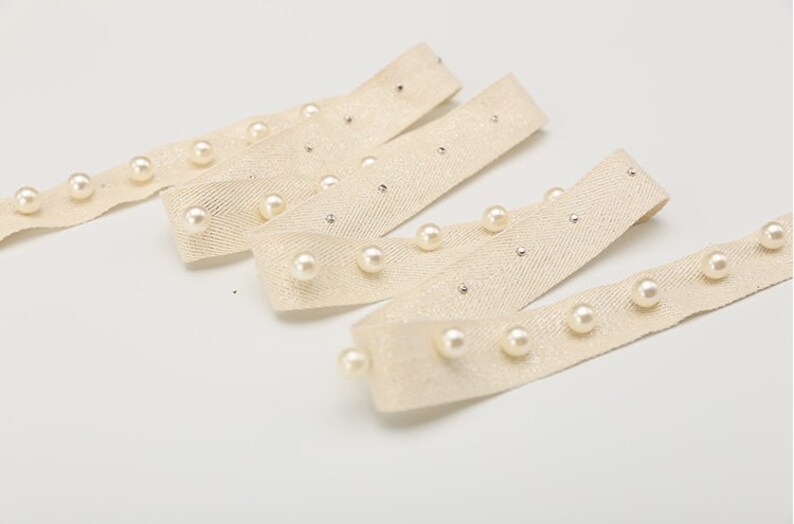 May include: Beige ribbon trim with white pearl embellishments. The ribbon has a herringbone weave and is decorated with evenly spaced pearls.