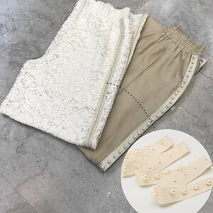 May include: Two pairs of pants, one white lace and one beige with pearl trim. A close-up of beige trim with pearls.