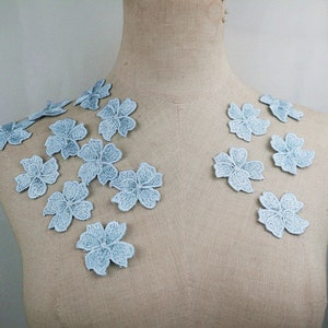 Pode incluir: Uma coleção de aplicações de flores de renda azul claro. As aplicações são dispostas sobre uma superfície de tecido marrom claro.