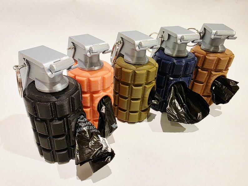 May include: A collection of five dog waste bag dispensers shaped like hand grenades. Each dispenser is a different color: black, coral, olive green, navy blue, and brown. The top and pull pin are silver, and a black bag is visible in the opening.