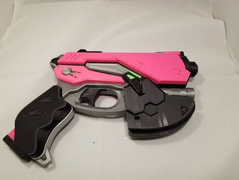 Puede incluir: Una pistola de juguete de pl&aacute;stico rosa y negro con un ca&ntilde;&oacute;n plateado y un detalle verde. La pistola tiene un agarre texturizado y un cargador extra&iacute;ble.