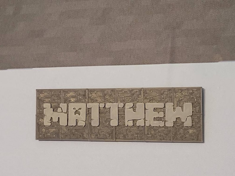 Pode incluir: Uma placa de nome bege com a palavra "MATTHEW" escrita em letras em relevo. As letras s&atilde;o feitas de um material texturizado que se assemelha a pequenos blocos.