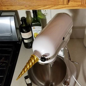 May include: A white stand mixer with a gold unicorn horn attachment. The mixer has a silver bowl and is sitting on a countertop.