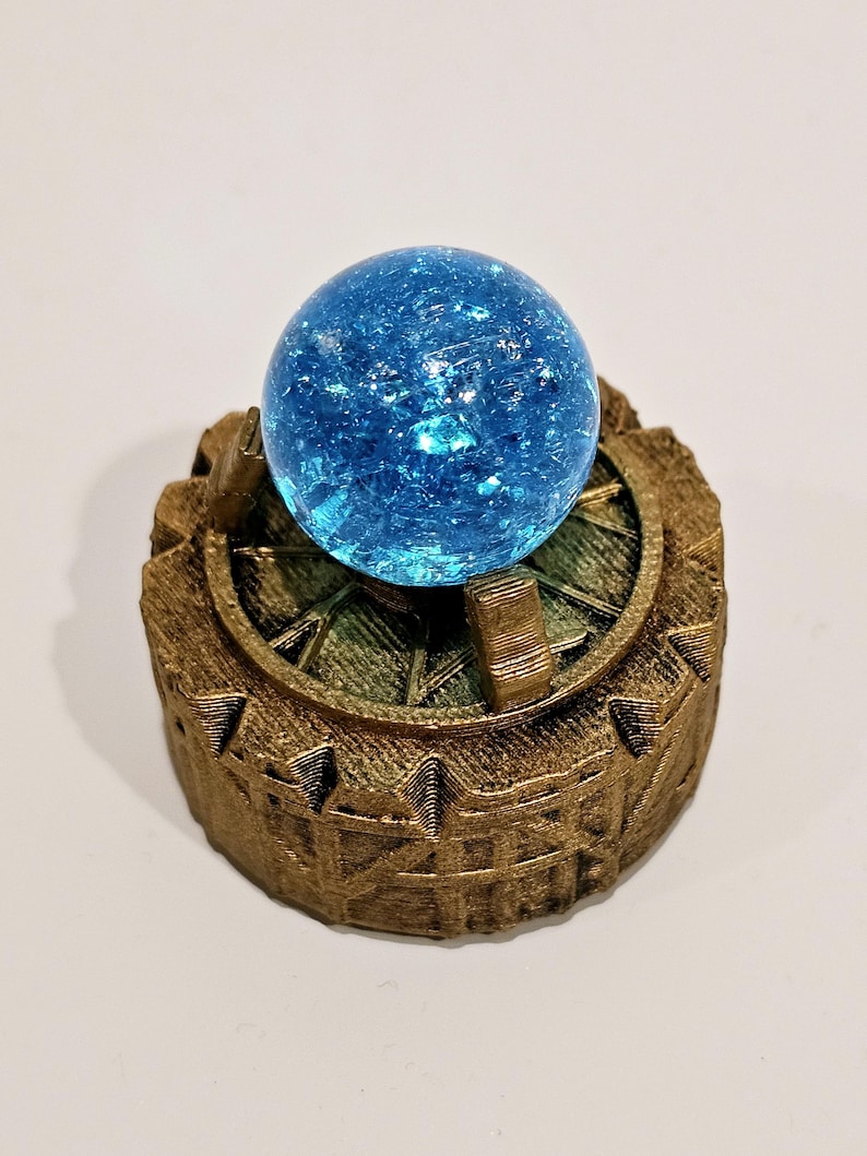 May include: A bright blue crackle glass sphere sits centred atop a circular, textured bronze-coloured base. The base features a raised, gear-like rim with vertical notches and a detailed, grid-patterned surface surrounding the central blue orb.