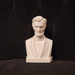 May include: White bust sculpture of Abraham Lincoln, a historical figure, with a serious expression. The bust is made of a smooth, white material and is set on a small, square base.