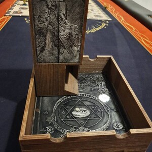 May include: A wooden dice tower with a black and white image of a creature on the front. The tower has a clear plastic tray with a black and white design on the bottom. The tray has a small white die in the center.