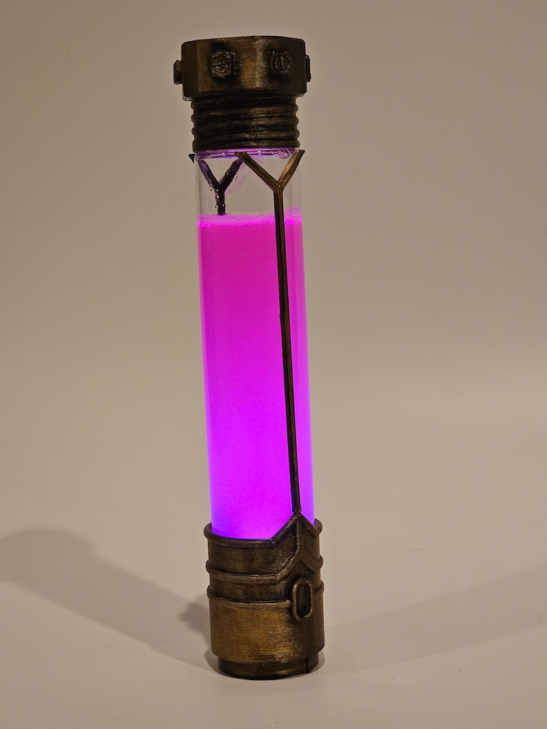 May include: A glass tube with a pink liquid inside, encased in a gold-colored metal frame. The tube has a decorative top and bottom.