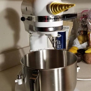 May include: A white KitchenAid stand mixer with a silver bowl and a gold-colored attachment. The mixer is on a white countertop.