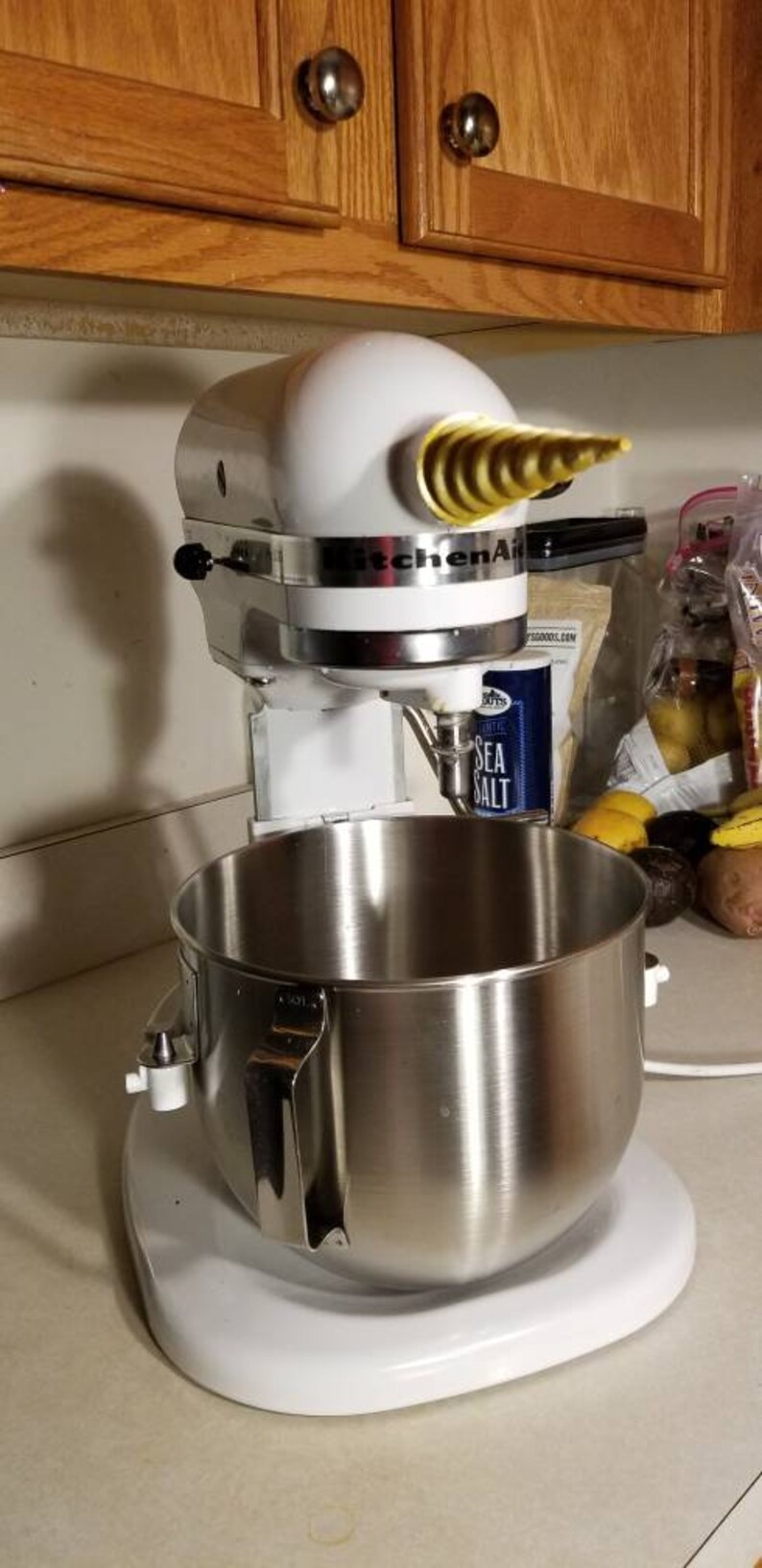May include: A white KitchenAid stand mixer with a silver bowl and a gold-colored attachment. The mixer is on a white countertop.