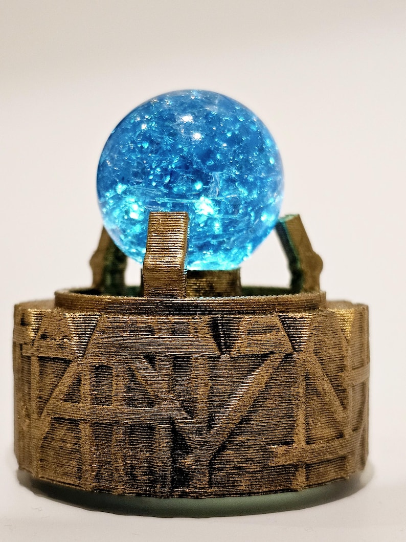 May include: A bright blue crackle glass sphere resting on a circular bronze-coloured base. The base features a textured, layered surface with geometric, angular carvings and three vertical prongs that hold the glowing blue orb in place.