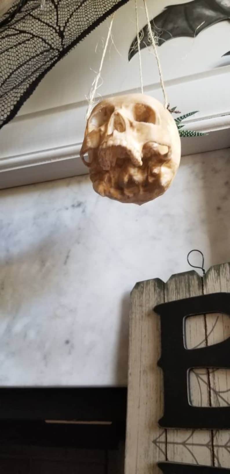 May include: A white plastic skull hanging from a string. The skull is hanging in front of a white wall with a black lace overlay.