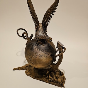 May include: A gold-colored eagle sculpture with its wings spread wide, perched atop a globe with a rope-wrapped anchor. The globe is resting on a textured base.